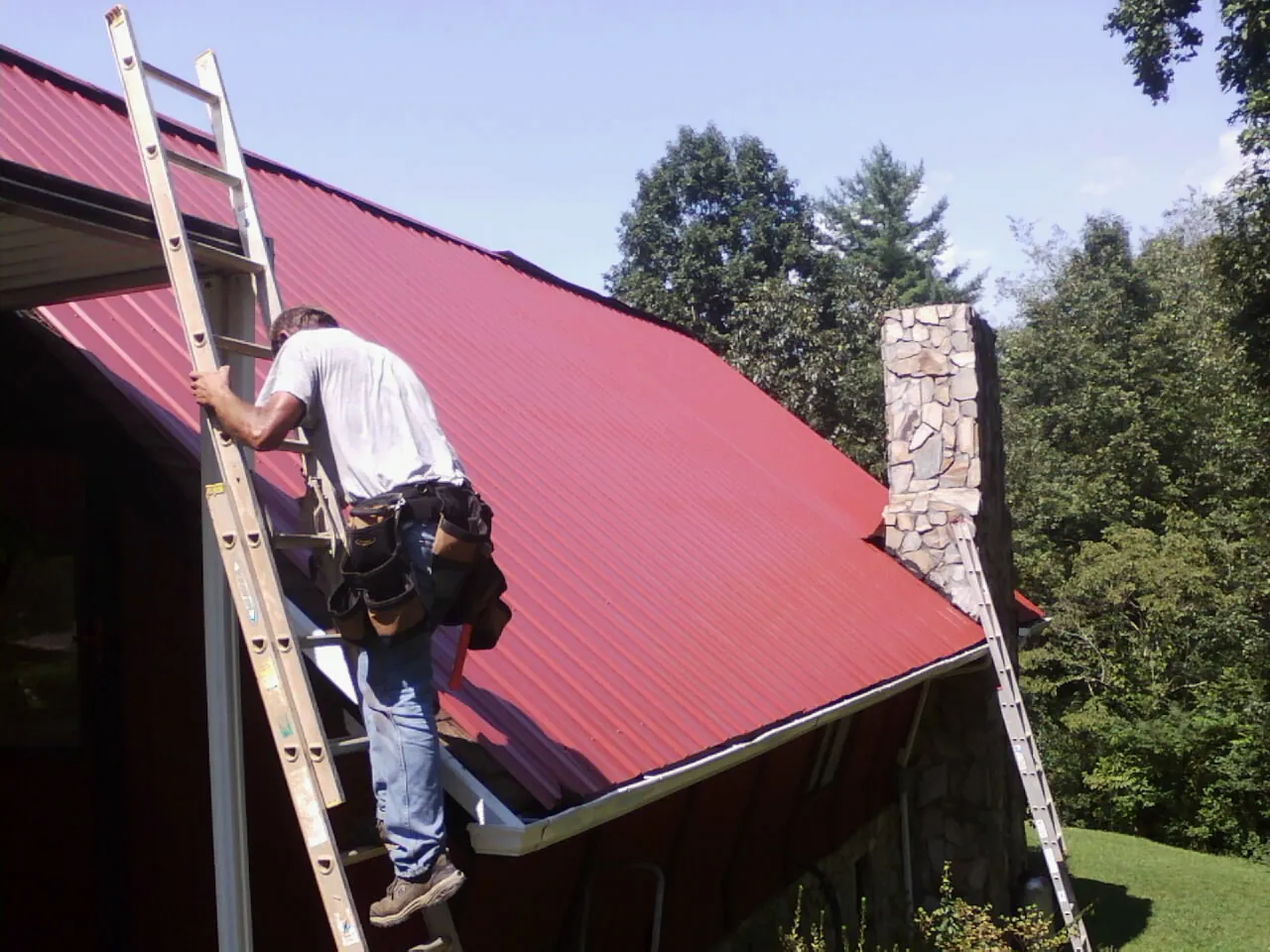 Roofer on a ladder beside a red metal roof and stone chimney — Soffit & Fascia Repair by Roofing Brothers in Marcola
